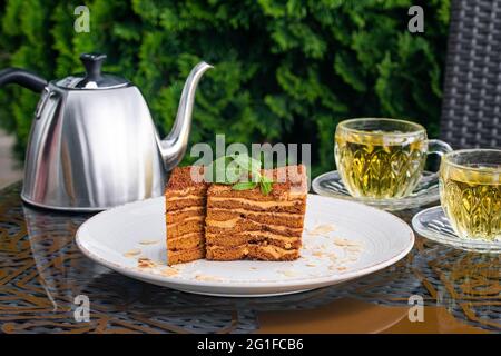 Gâteau Medovik recouvert de chapelure, deux tasses de thé à la menthe et bouilloire en acier sur table dans le café d'été. Concept de boisson au thé, culture britannique. Encore la vie. Banque D'Images