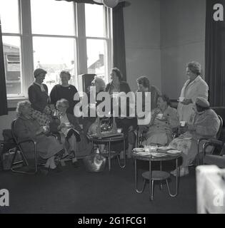 Années 1960, historique, à l'intérieur d'une salle de séjour dans une maison de soins, un groupe de femmes âgées assis dans des chaises autour d'une théière en métal, sur un stand en bois, enfermé « The Singing Kettle Club », étant servi thé et biscuits par le personnel féminin, Écosse, Royaume-Uni. Banque D'Images