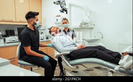 Homme caucasien assis dans une chaise de dentiste discutant avec l'infirmière mâle et femelle au sujet de l'opération dentaire Banque D'Images