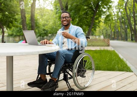 Travail à distance pour les personnes handicapées. Joyeux homme noir en fauteuil roulant travaillant en ligne, en utilisant un ordinateur portable au café extérieur Banque D'Images