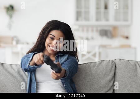 Femme noire ludique utilisant le joystick pour jouer à des jeux vidéo en ligne, assis sur un canapé dans le salon, espace vide Banque D'Images