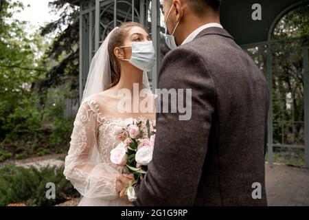 mariée dans le masque médical regardant le marié tout en tenant le bouquet à l'extérieur Banque D'Images