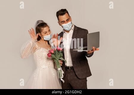 mariée et marié dans des masques médicaux agitant les mains pendant l'appel vidéo isolé sur gris Banque D'Images