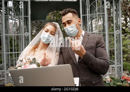 nouveaux mariés dans des masques de protection montrant des anneaux de mariage pendant le chat vidéo sur ordinateur portable Banque D'Images