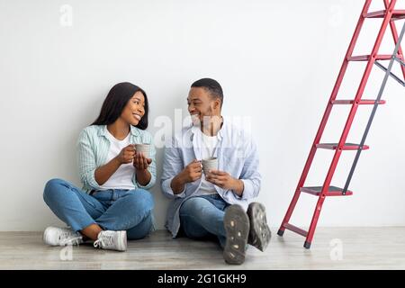 Un jeune couple afro-américain discute du plan de réparation dans sa nouvelle maison et de boire du café, assis sur le sol Banque D'Images
