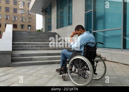 Un homme noir handicapé contrarié en fauteuil roulant devant les escaliers sans rampe, n'ayant pas la possibilité d'entrer dans le bâtiment Banque D'Images