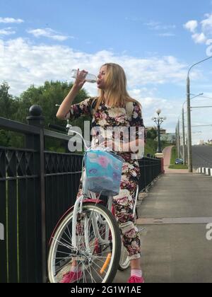 Une fille dans un survêtement posant sur un vélo. Boisson de l'eau d'une bouteille en plastique. Se trouve près des rails du pont routier. Banque D'Images