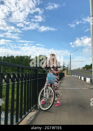 Une fille dans un survêtement posant sur un vélo. Boisson de l'eau d'une bouteille en plastique. Se trouve près des rails du pont routier. Banque D'Images