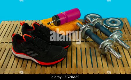 Ensemble de gym avec paire de chaussures de sport noires, haltères pour l'exercice, serviette jaune et bouteille rouge avec eau sur parquet avec fond bleu Banque D'Images