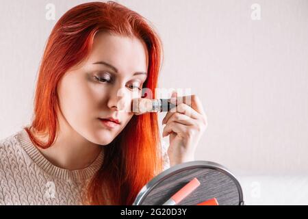 Jeune femme à tête rouge regarde dans le miroir et applique le maquillage à son visage. Les femmes utilisent des cosmétiques. Banque D'Images