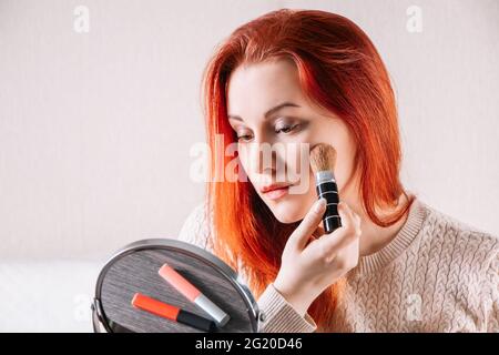 Jeune femme à tête rouge regarde dans le miroir et applique le maquillage à son visage. Les femmes utilisent des cosmétiques. Banque D'Images