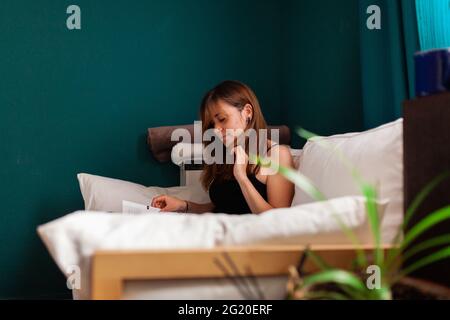 Jeune femme aux cheveux rouges, lisant un livre dans un canapé entouré de plantes. Loisirs en intérieur. Banque D'Images