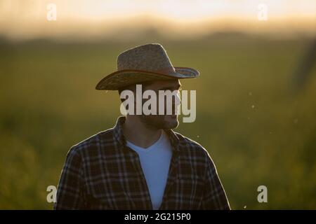 Portrait d'un jeune agriculteur avec chapeau de paille debout au champ au coucher du soleil au printemps Banque D'Images