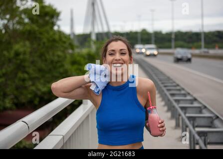 Jeune femme se reposant après l'entraînement essuyant la sueur souriant. L'athlète hydratant ayant Milk-Shake de fraise sur le pont. Banque D'Images