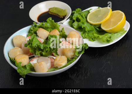 Viande de pétoncle frite aux crevettes bouillies dans une assiette en céramique avec laitue, citron, sauce soja et wasabi sur fond sombre. Mise au point sélective. Banque D'Images