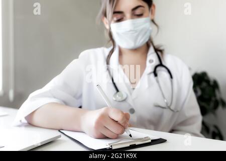 Le médecin en robe de shite avec stéthoscope et masque de protection du visage écrit quelque chose dans les antécédents médicaux du patient. Photo de haute qualité Banque D'Images