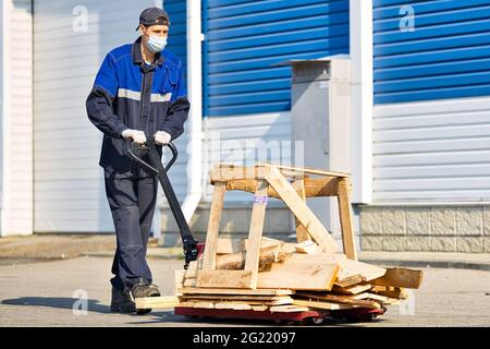 Un homme en vêtements de travail et un masque médical porte des planches en bois sur une charrette sur le territoire d'une base industrielle pendant une journée d'été. Travailler conformément aux normes sanitaires Banque D'Images