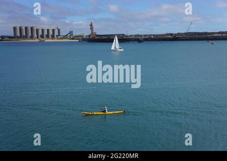 Un kayak jaune et un voilier se dirigeant dans les eaux bleues de Mackay, Queensland, dans le port, sur fond de quais commerciaux et de silos à grains. Banque D'Images