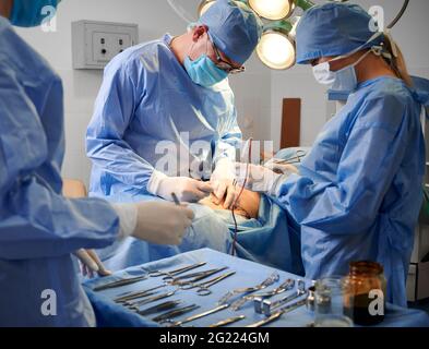 Équipe chirurgicale de gants stériles et de masques de protection en chirurgie plastique dans la salle d'opération. Médecins effectuant la chirurgie esthétique tout en assistant debout près de la table chirurgicale avec des instruments médicaux. Banque D'Images