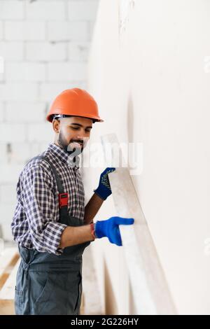 Un ouvrier en chapeau, tenant un couteau à mastiquer à peigne et couvrant une surface de mur avec de la pâte adhésive avant de poser la pierre décorative. Travaux de rénovation et Banque D'Images