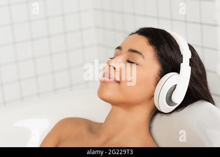 jeune femme souriante avec les yeux fermés se reposant et écoutant de la musique dans les écouteurs de la salle de bains Banque D'Images