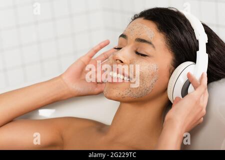 bonne femme afro-américaine avec un gommage sur le visage allongé dans la baignoire avec les yeux fermés et de la musique dans les écouteurs Banque D'Images