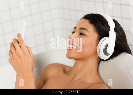 jeune femme souriante écoutant de la musique dans un casque regardant un téléphone portable dans la salle de bains Banque D'Images