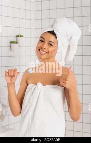 jeune femme afro-américaine souriante enveloppée dans des serviettes tenant la brosse à dents et montrant un geste ok dans la salle de bains Banque D'Images