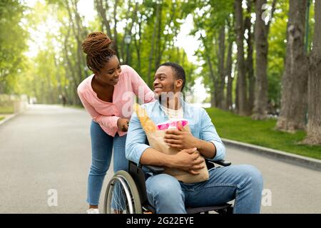 Joyeux jeune homme handicapé en fauteuil roulant transportant un sac en papier avec de la nourriture, sa belle femme l'aidant à l'extérieur Banque D'Images