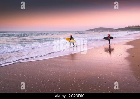 Coucher de soleil sur les surfeurs marchant dans la mer à Fistral Beach à Newquay dans les Cornouailles. Banque D'Images