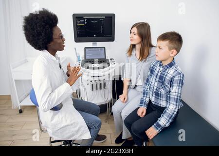 Jeune femme de 30 ans de race blanche avec son fils adolescent médecin traitant dans la salle d'examen moderne d'échographie. Femme afro-américaine médecin expliquant la procédure d'échographie à ses patients Banque D'Images