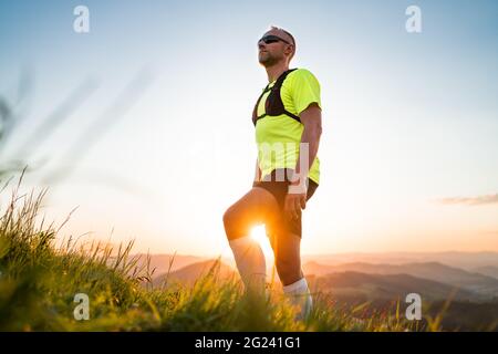 Portrait d'Active Mountain Trail Runner habillé t-shirt brillant avec sac à dos et lunettes de soleil de sport de repos pendant qu'il court le ciel par les collines pittoresques a Banque D'Images