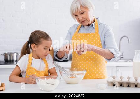 Femme âgée versant des œufs dans un bol près des ingrédients et petite-fille dans la cuisine Banque D'Images
