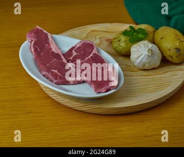 Steak de bœuf cru avec pototes crus, gants à l'ail et feuilles de persil sur une assiette ronde en bois Banque D'Images