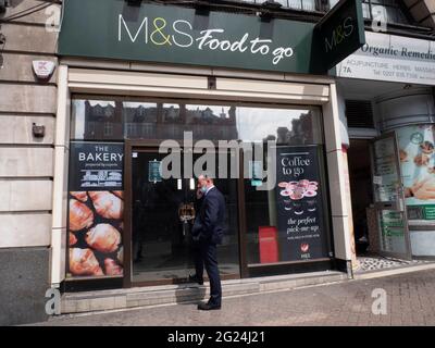 Sous clé et fermé Marks and spencer food à entreposer avec un homme portant un masque de coronavirus sur le téléphone debout dans la porte Banque D'Images