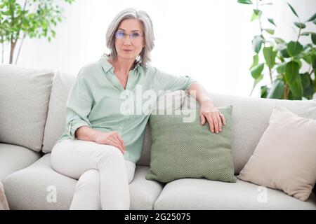 Photo de charmante sérieuse calme vieille femme s'asseoir canapé week-end porte des lunettes à l'intérieur de la maison appartement Banque D'Images