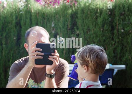Père prenant une photo de son fils tenant une machine à bulles avec une tablette numérique Banque D'Images