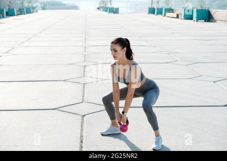 Brunette focalisée s'exerçant sur la chaussée à l'extérieur avec peu de kettlebell, le balançant vers l'avant de la position de squat. Haut gris et pantalon de yoga. Banque D'Images