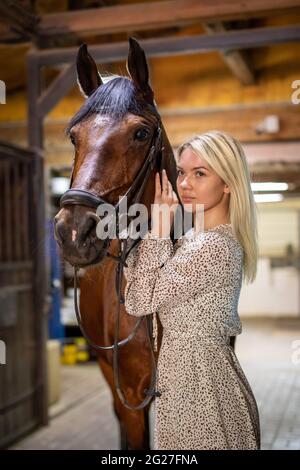 Une jeune rider femme blonde avec de longs cheveux dans une robe posant avec un cheval brun à l'intérieur stable, Sibérie, Russie Banque D'Images