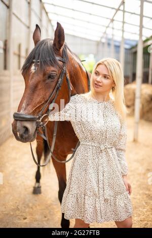 Une jeune rider femme blonde avec de longs cheveux dans une robe posant avec un cheval brun à l'intérieur de lumière stable, Sibérie, Russie Banque D'Images