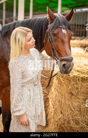 Une jeune rider femme blonde avec de longs cheveux dans une robe posant avec un cheval brun à l'intérieur de lumière stable, Sibérie, Russie Banque D'Images