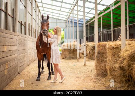 Une jeune rider femme blonde avec de longs cheveux dans une robe posant avec un cheval brun à l'intérieur de lumière stable, Sibérie, Russie Banque D'Images