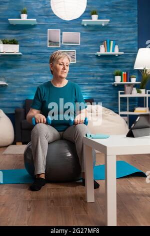 Femme âgée exerçant des muscles du corps faisant des bras exercice assis sur le ballon suisse dans le salon. Pensioner regardant la formation en ligne d'aérobic sur le comprimé d'exercice de résistance musculaire tenant dumbbell Banque D'Images