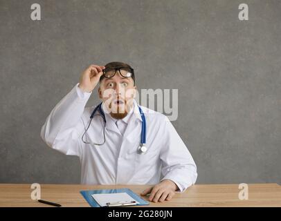 Un médecin choqué, un professionnel de la santé, prend des lunettes, s'assoit au bureau et regarde la caméra Banque D'Images