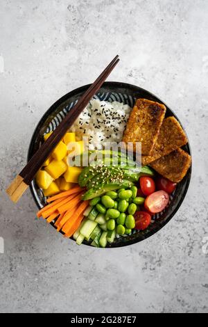 Vue de haut en bas d'un bol de poke vegan maison avec tofu, mangue, avocat, carotte, edamame, tomate, concombre sur fond gris, verticale Banque D'Images