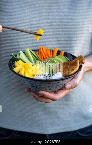 Une femelle dans une chemise bleue tenant un morceau de mangue avec des baguettes au-dessus d'un bol de poke frais avec de la mangue, de l'avocat et du tofu, bol vegan, vertical Banque D'Images