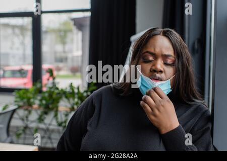 femme d'affaires afro-américaine de plus grande taille qui prend son masque médical tout en se sentant mal au bureau Banque D'Images