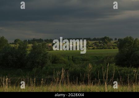 Un village entouré de verdure sur les rives de la rivière, sur fond de ciel sombre et orageux. Paysage rural avec maisons de village. Banque D'Images