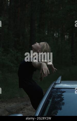 Une belle fille blonde en vêtements noirs est debout en se penchant hors de la fenêtre de voiture, dans une posture gracieuse avec les bras étirés et la tête projetée en arrière. Banque D'Images