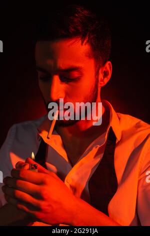 Homme barbu avec une cigarette tenant l'allume-cigare en lumière rouge sur fond noir Banque D'Images